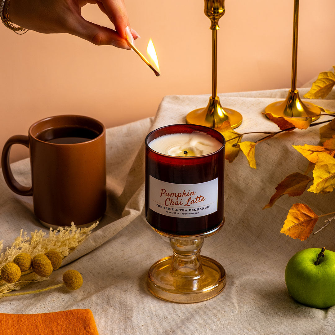 A lit candle in a glass pedestal holder on a draped fabric surface, with a hand holding a match near the flame. A brown mug, gold candlesticks, yellow leaves, and a green apple surround it in a styled autumn scene.